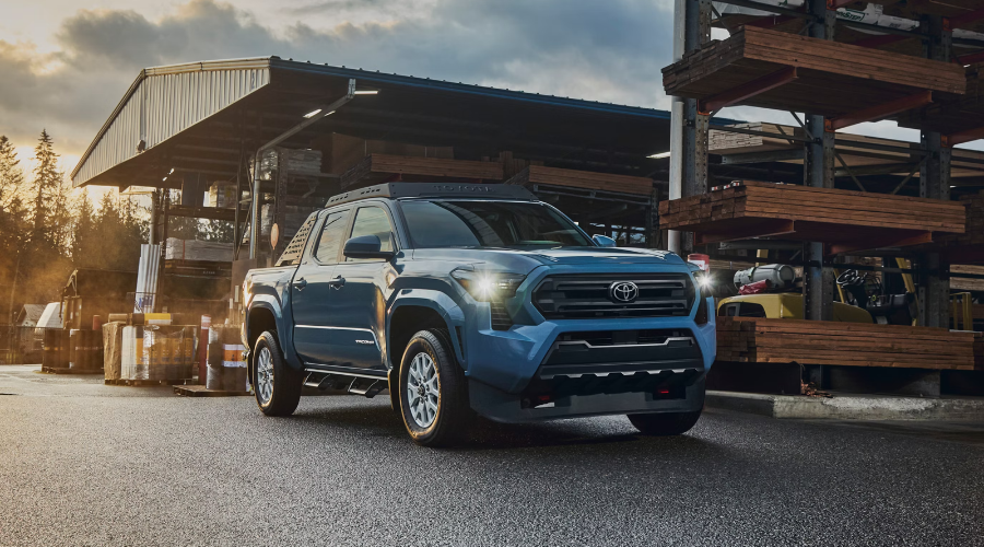 Front view of a blue 2026 Toyota Tacoma 4WD midsize pickup truck with headlights on parked at a damp Lake County industrial job site, demonstrating wet weather driving capability in Waukegan, IL.